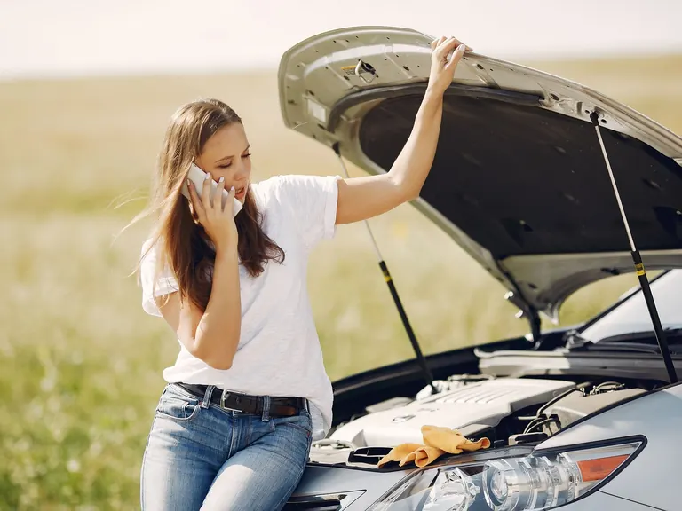 Conductora habla por celular apoyada en su auto con el capó abierto en una carretera rural